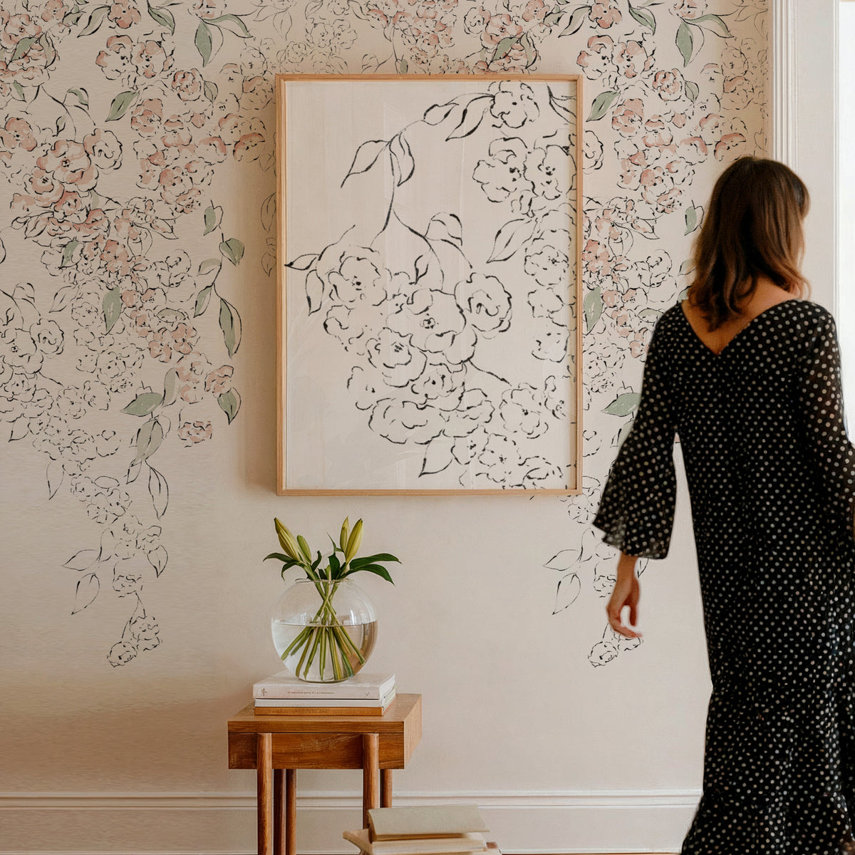 A woman in a black polka-dot dress stands in a room with floral wallpaper, looking at a large framed black line-art floral print on the wall. A small wooden side table with a glass vase of lilies sits below the frame.