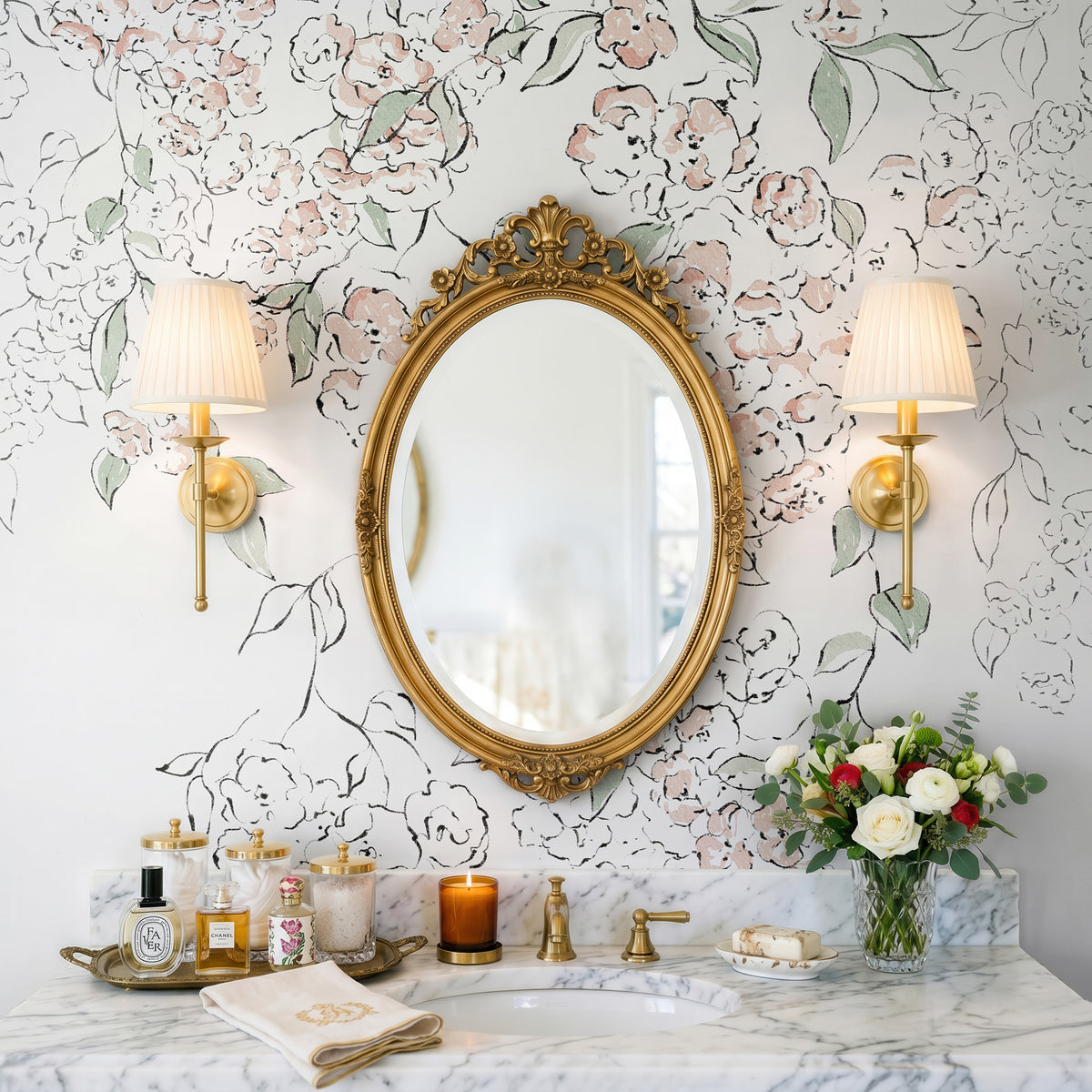 An ornate oval gold-framed mirror hangs on a white floral wallpapered wall above a marble bathroom vanity with gold fixtures, two lit wall sconces, toiletries, and a vase of flowers.