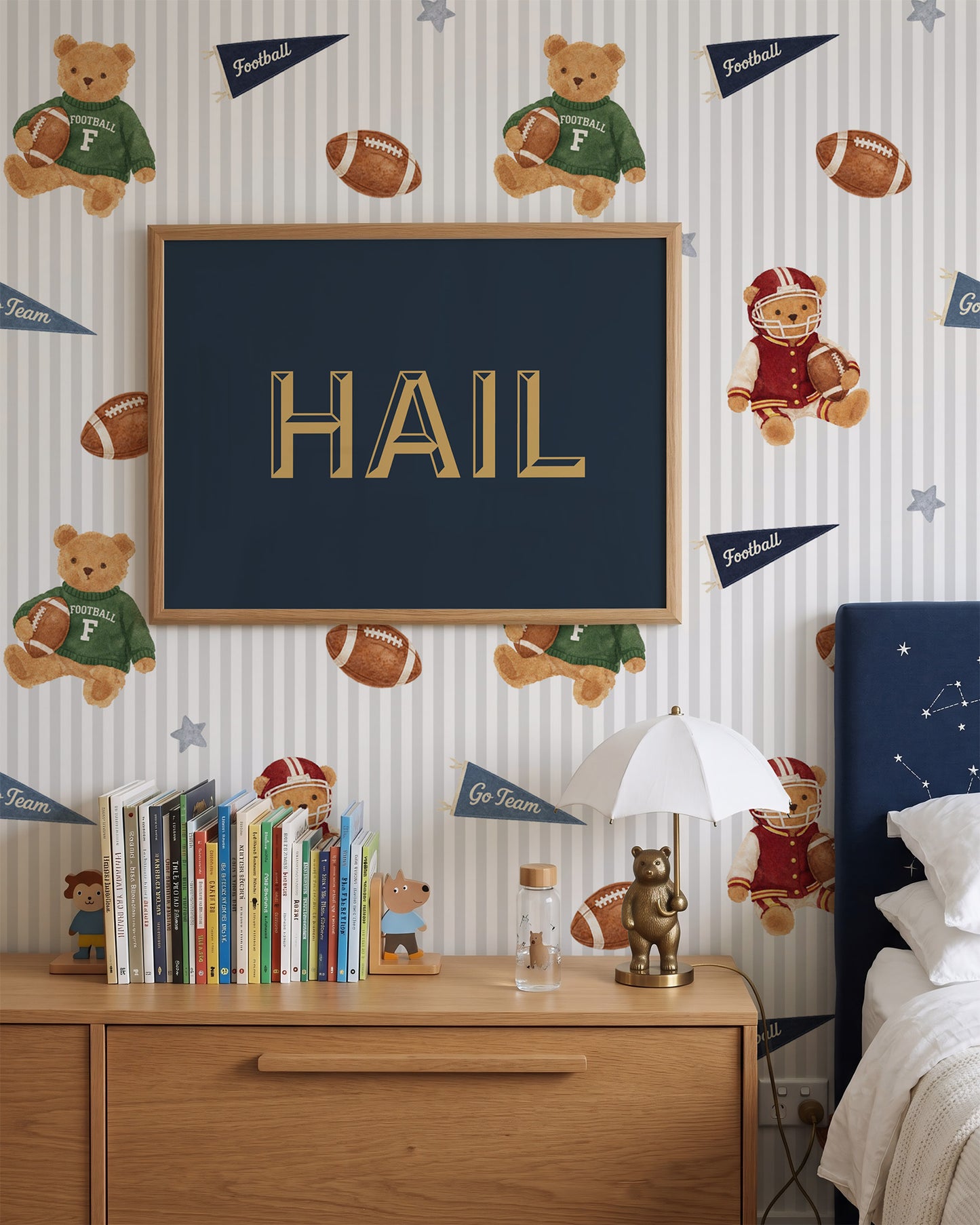 A styled children's bedroom featuring a custom framed 'HAIL' sign with gold lettering on a navy blue background, mounted on a striped wallpaper decorated with football-themed teddy bears, footballs, and pennants. Below the sign is a wooden dresser with books, a small lamp, and a glass bottle, next to a dark blue upholstered bed headboard.