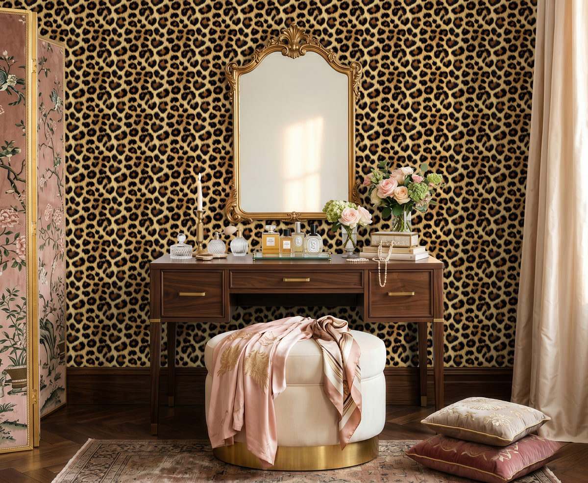 A styled interior scene featuring a dark wood vanity table with a large ornate gold-framed mirror, set against a bold leopard-print wallpaper. A round cream ottoman with a gold base and draped pink satin sits in front, with decorative flowers, perfume bottles, and pillows on and around it.