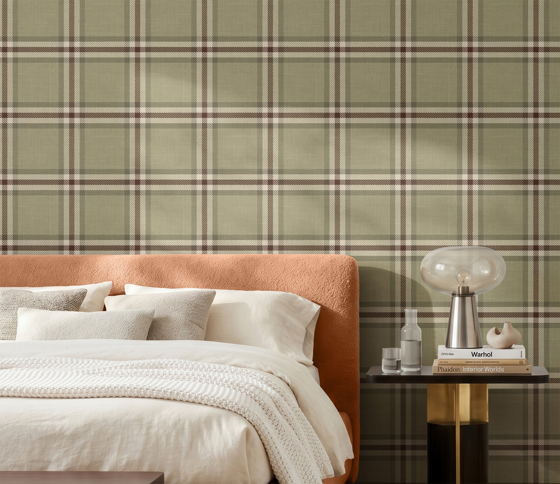 A styled bedroom scene featuring a bed with white bedding and a textured cream throw, set against a wall with sage green plaid wallpaper. A modern orange upholstered headboard is visible on the left, and a dark side table with a glass table lamp and books sits on the right.