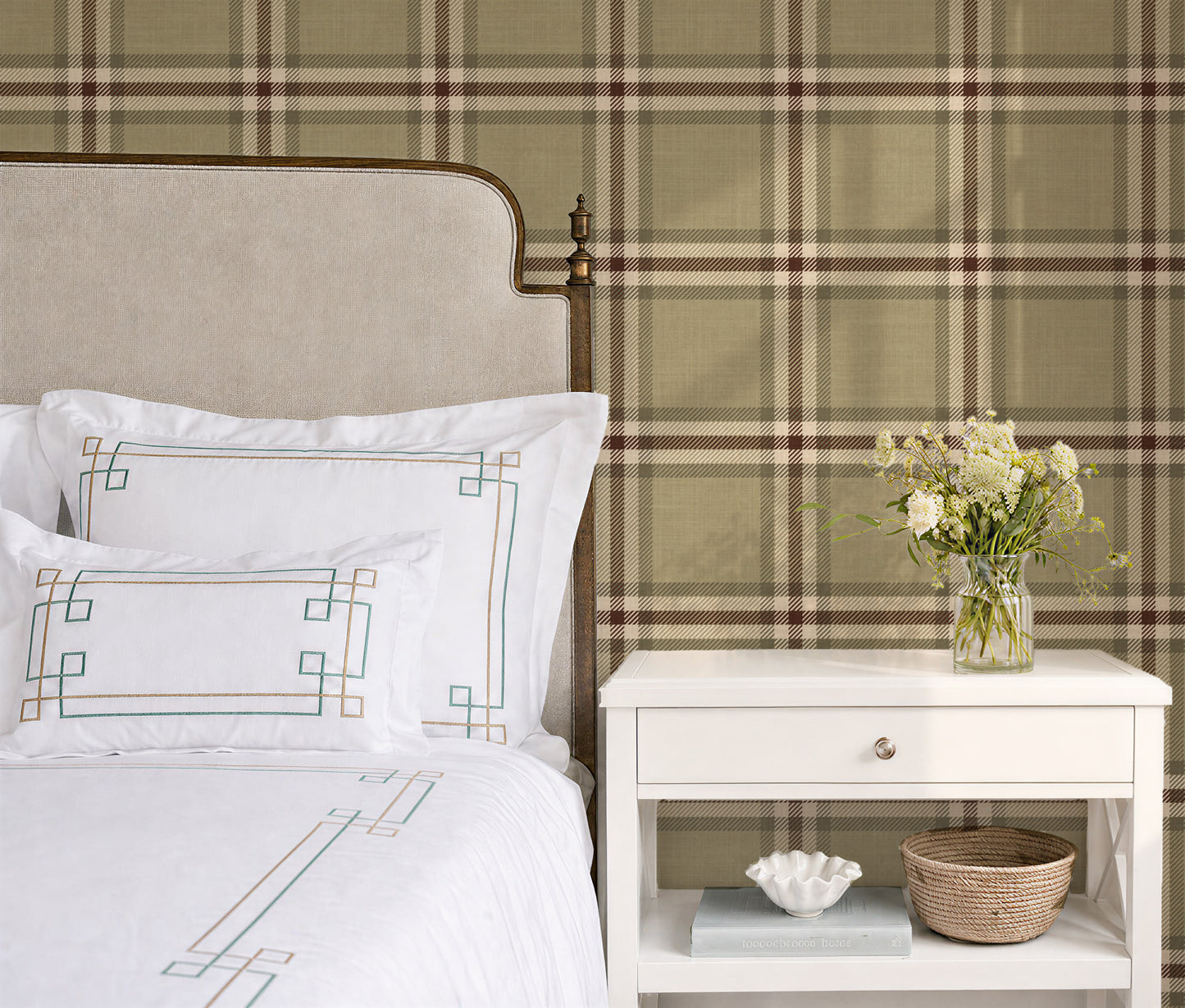 A styled bedroom corner featuring a bed with white linens and embroidered pillows, a white nightstand with a vase of flowers, and a large plaid wallpaper backdrop in muted green, beige, and brown tones.