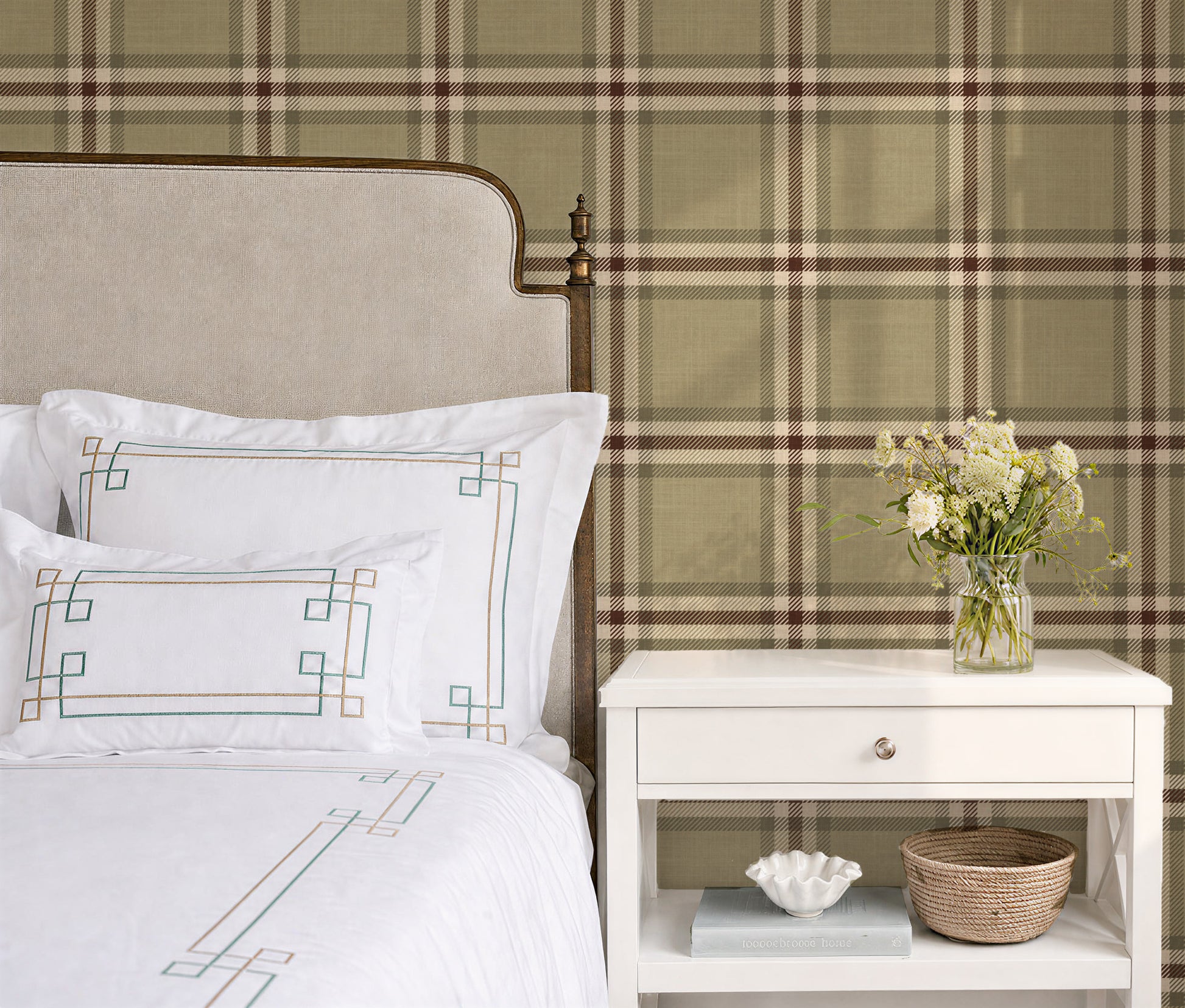 A styled bedroom corner featuring a bed with white linens and embroidered pillows, a white nightstand with a vase of flowers, and a large plaid wallpaper backdrop in muted green, beige, and brown tones.